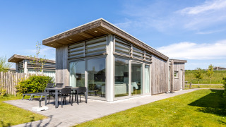 Moderne Lodge 4 med træfacade, store vinduer og terrasse med havemøbler på en solrig dag.