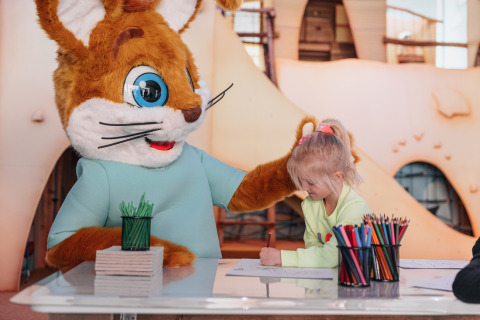 Un niño dibuja en una mesa con lápices de colores junto a una mascota gigante de conejo en un parque cubierto.
