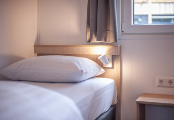 Modernes Bett mit weißer Bettwäsche, Leselampe und Fenster in Timber Cottage 4, Bospark 't Wolfsven, Niederlande.