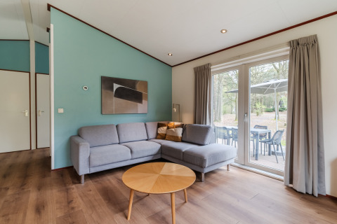 Stue i chalet WVR6 på Bospark 't Wolfsven med grå hjørnesofa, træbord og udsigt til terrasse.