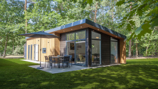 Moderne Lodge 4 tiny house i skoven med store vinduer, terrasse, parasol og havemøbler på græsplæne.