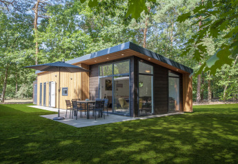 Moderne Lodge 4 tiny house i skoven med store vinduer, terrasse, parasol og havemøbler på græsplæne.