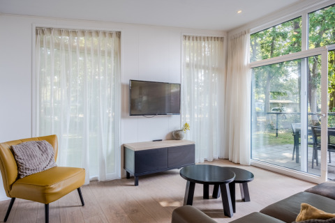 Bright living room with yellow chair, TV, large windows, and patio view in Lodge 4 at Bospark 't Wolfsven.