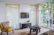 Lys stue med gule og sorte møbler, tv, store vinduer og udsigt til terrasse i Bospark 't Wolfsven.