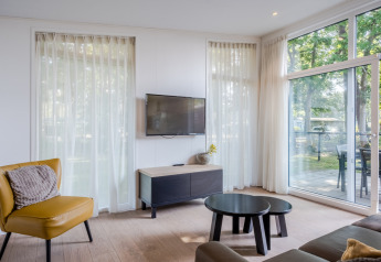 Lys stue med gule og sorte møbler, tv, store vinduer og udsigt til terrasse i Bospark 't Wolfsven.