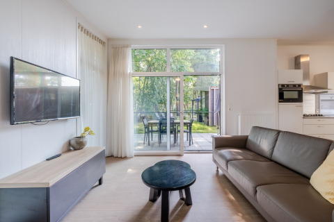 Bright living area in Lodge 4 at Bospark 't Wolfsven, Netherlands, with sofa, TV, patio and garden view.