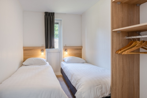 Pequeña habitación en una tiny house con dos camas individuales, ventana, cortina y armario abierto con perchas.