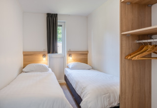 Piccola camera da letto in una tiny house con due letti singoli, finestra, tenda e armadio aperto con grucce.