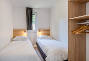 Kleines Schlafzimmer in einem Tiny House mit zwei Einzelbetten, Fenster, Vorhang und offenem Kleiderschrank.
