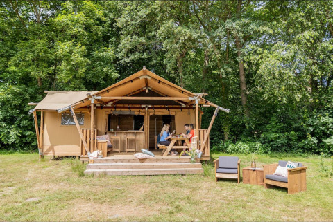 Ein gemütliches Safarizelt mit einer Veranda, auf der Menschen an einem Tisch sitzen, umgeben von Bäumen.