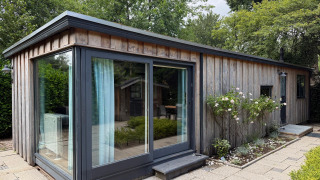 Modernes Holzhaus mit großen Glasfenstern, umgeben von Blumen und Bäumen in Het Verscholen Dorp, Niederlande.