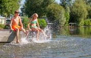 Zwei Kinder in Badekleidung sitzen auf einem Steg im Ferienpark Tolplas, Overijssel, Niederlande und planschen mit den Füßen im Wasser.