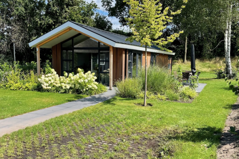 Modernes Schipbeek 6 Lodge mit großen Fenstern, umgeben von Garten und Natur im Sonnenschein.