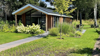 Modernes Schipbeek 6 Lodge mit großen Fenstern, umgeben von Garten und Natur im Sonnenschein.