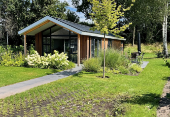 Lodge Schipbeek 6 moderne en bois avec grandes fenêtres, entouré d’un jardin verdoyant sous le soleil.