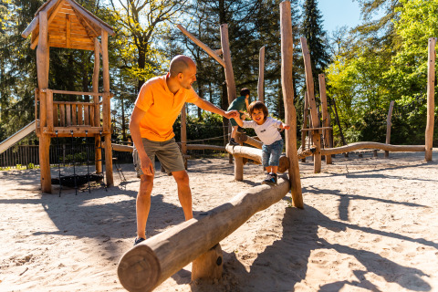 Man helpt kind balanceren op houten boomstam in een speeltuin aan een lodge midden in het bos.