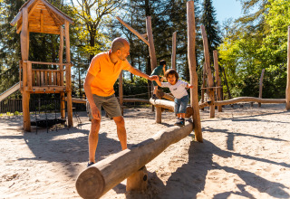 Man helpt kind balanceren op houten boomstam in een speeltuin aan een lodge midden in het bos.
