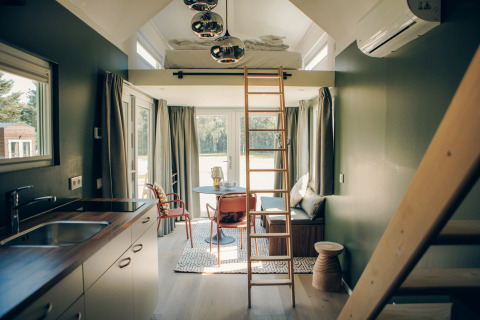 Modern, sunlit interior of Koya Tiny Loft tiny house in Germany, featuring loft bed, ladder, kitchen, and sitting area.