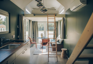 Modern, sunlit interior of Koya Tiny Loft tiny house in Germany, featuring loft bed, ladder, kitchen, and sitting area.