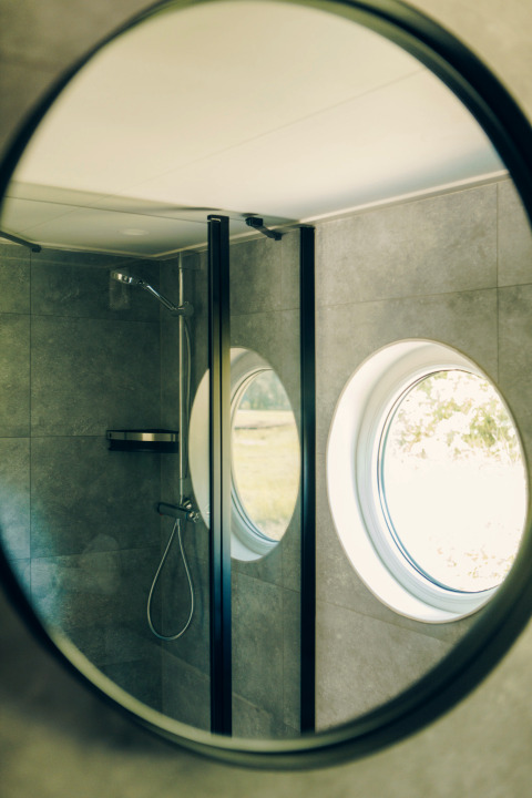 Round mirror reflecting a small bathroom with shower and circular window at Koya Tiny Loft, Wilsumer Berge.