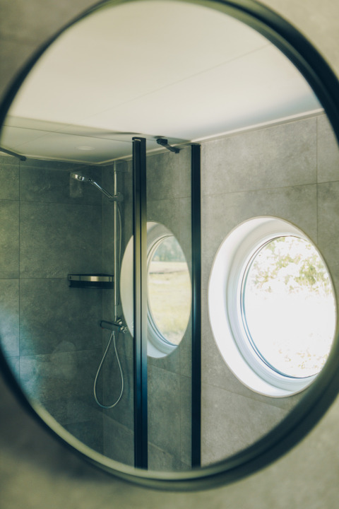 Round mirror reflecting a small bathroom with shower and circular window at Koya Tiny Loft, Wilsumer Berge.