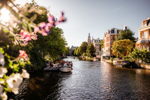 Vista soleada de un canal con flores, gente en un barco y edificios históricos en un parque de glamping.
