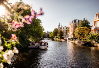 Solrig kanaludsigt med blomster, folk på båd og historiske bygninger ved et feriepark med glamping.