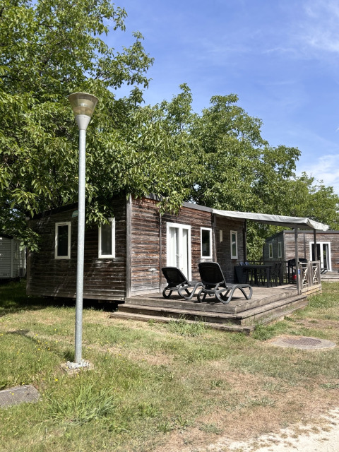 Buitenzicht op Mobile Home Premium met houten veranda, ligzetels en groene bomen rondom.