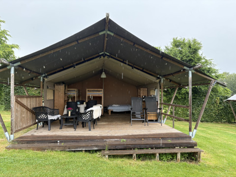 Safari tent with wooden deck, garden chairs, and open front, surrounded by green lawn on a rainy day.