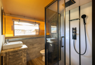 Baño dentro de la tienda Safari 6 en Hunzepark, Países Bajos, con ducha, lavabo, madera y ventana amplia.