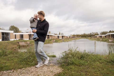 En mand bærer et lille barn ved en sø foran moderne glamping-hytter under en overskyet himmel.