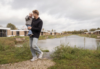 Hombre con un niño en brazos junto a un estanque y frente a modernas cabañas de glamping en día nublado.