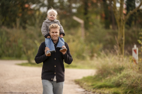 En mand går udenfor med et smil, mens et lille barn sidder på hans skuldre i naturlige omgivelser ved en lodge.