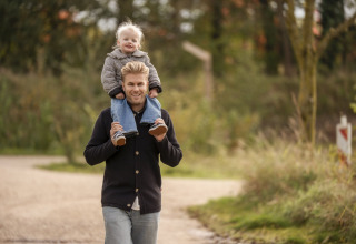 En mand går udenfor med et smil, mens et lille barn sidder på hans skuldre i naturlige omgivelser ved en lodge.