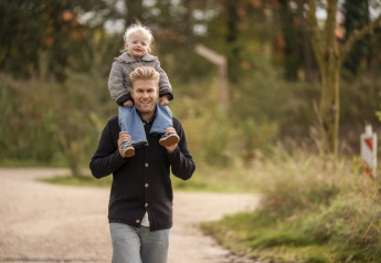 Een lachende man wandelt buiten met een kind op zijn schouders, omgeven door groen bij een lodge.