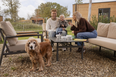 En familie sidder udenfor på et glampingsted, nyder kaffen sammen med deres barn og hund.