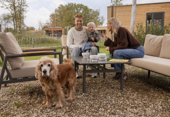 Een gezin zit buiten bij een glampingaccommodatie, genietend van koffie met hun kind en hond erbij.