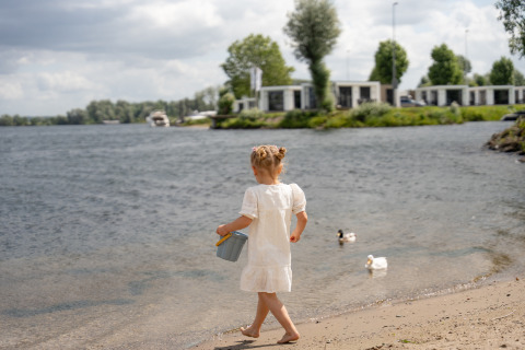 Kleines Mädchen im weißen Kleid spielt am Seeufer bei Villa Maritiem im MarinaPark Bad Nederrijn, Niederlande.