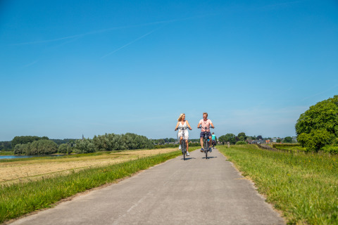 Couple cycling on a scenic path by a glamping accommodation under a clear blue summer sky.