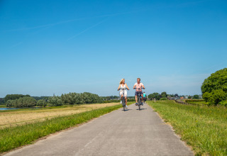 Couple cycling on a scenic path by a glamping accommodation under a clear blue summer sky.