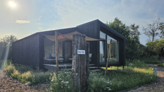 Moderne mørk træhytte ved Slowlodge 2 med skiltet 'Rise', solrig dag og vilde blomster i forgrunden.