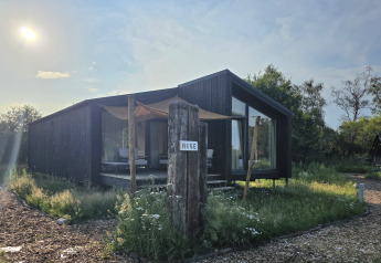 Moderne mørk træhytte ved Slowlodge 2 med skiltet 'Rise', solrig dag og vilde blomster i forgrunden.