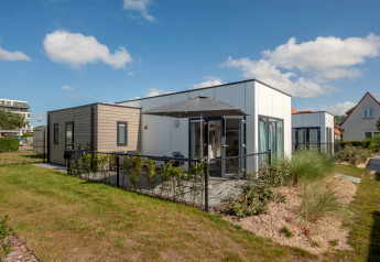 Moderne Lodge 4 mit Terrasse und schwarzem Zaun, umgeben von Pflanzen, unter blauem Himmel.
