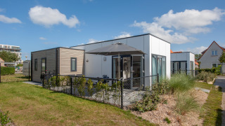 Moderne Lodge 4 med terrasse og sort hegn, omgivet af planter under en blå himmel med skyer.