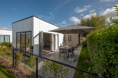Moderne Lodge 6 mit Terrasse, Gartenmöbeln und Sonnenschirm in einer grünen Umgebung bei Sonne.
