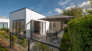 Moderne Lodge 6 med terrasse, havemøbler og parasol i et naturområde under en solrig himmel.