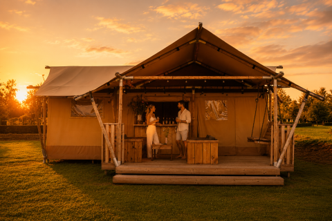 Ein luxuriöses Glamping-Zelt bei Sonnenuntergang, in dem zwei Personen auf einer Holzterrasse entspannen.