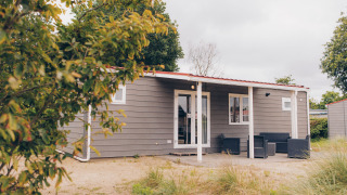 Außenansicht der Dune Lodge: Eine moderne, graue Hütte mit Veranda und Gartenmöbeln in natürlicher Umgebung.