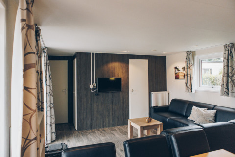 Modern living room in Dune Lodge at Duinhoeve, Netherlands, featuring black sofas, TV, and windows.