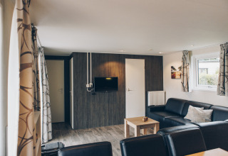Modern living room in Dune Lodge at Duinhoeve, Netherlands, featuring black sofas, TV, and windows.